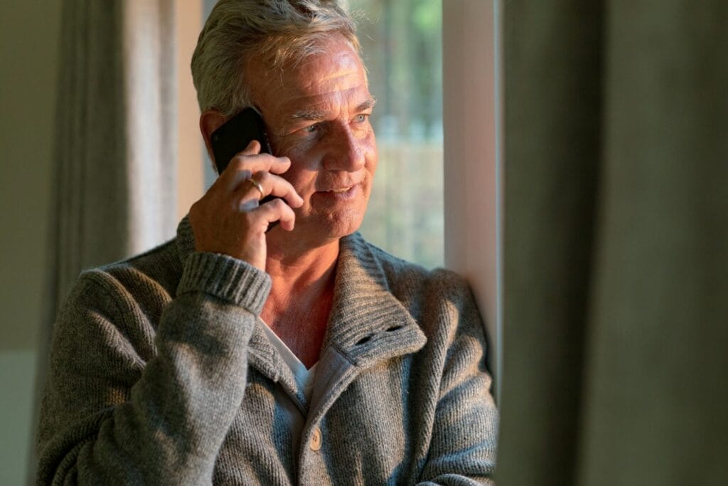 Happy caucasian senior man standing at window and making call