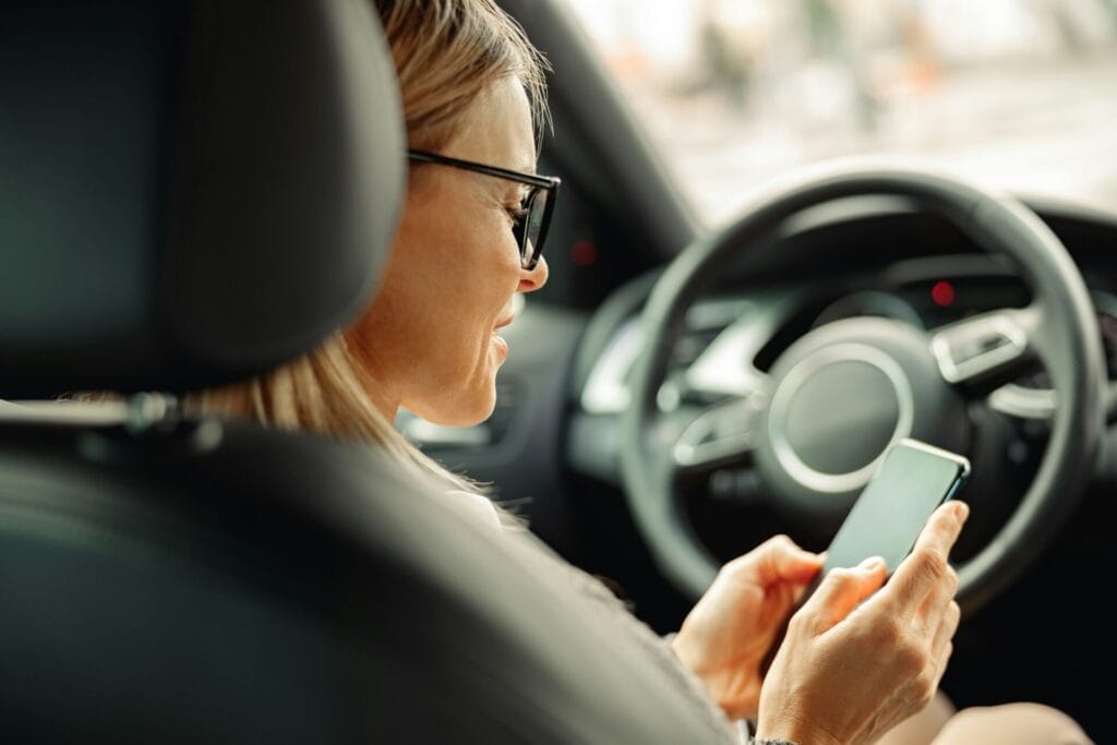 Side view of woman driver in eyeglasses is using phone while sitting behind steering wheel